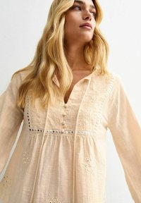 Helle beige Bluse mit gestickten Blumenmustern, Knopfdetail, silbernen Akzenten und lockerer Passform, ausgestattet mit einem Quast-Halsband und langen Ärmeln.