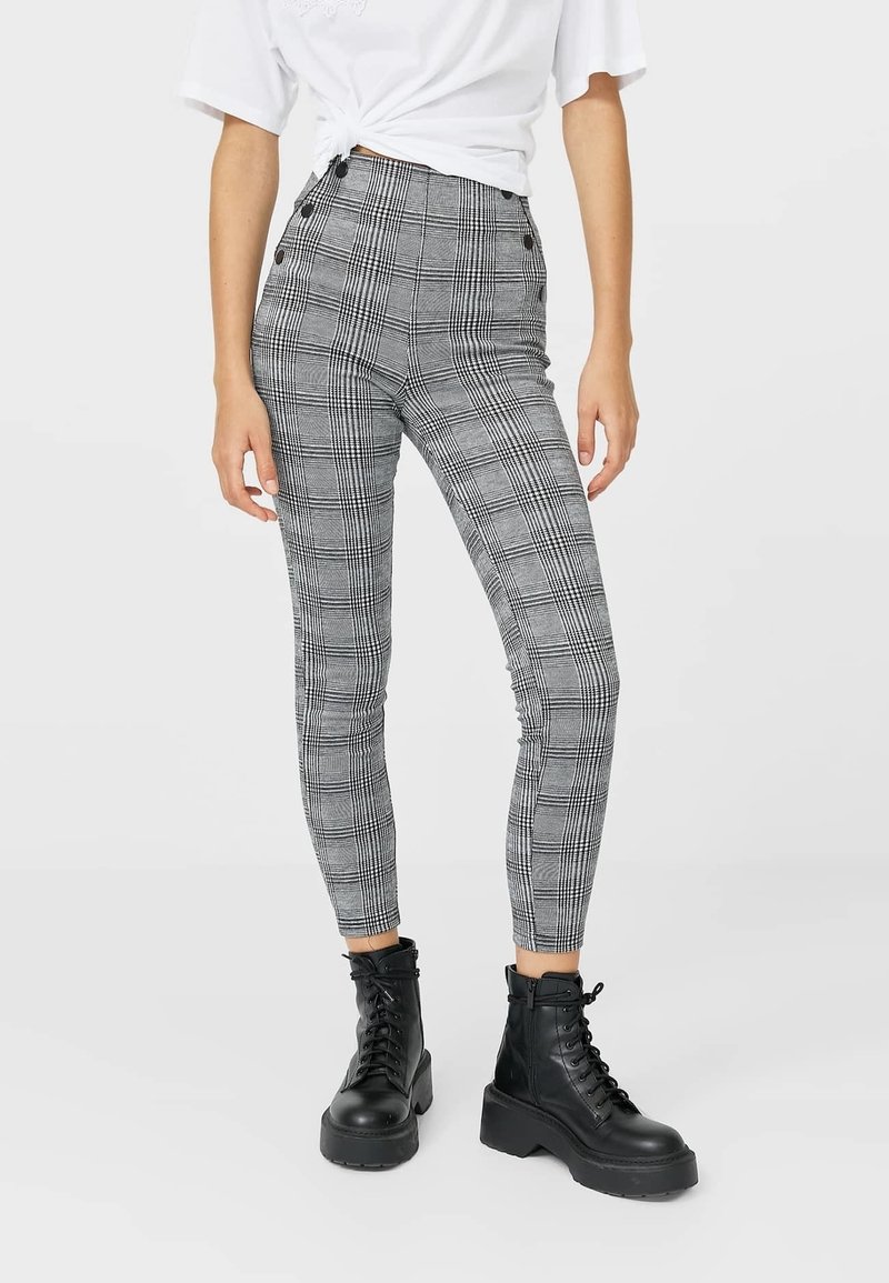 Pantaloni a vita alta, aderenti, a quadri in bianco e nero con motivo a scacchi. Presentano dettagli con bottoni in vita e una texture liscia.