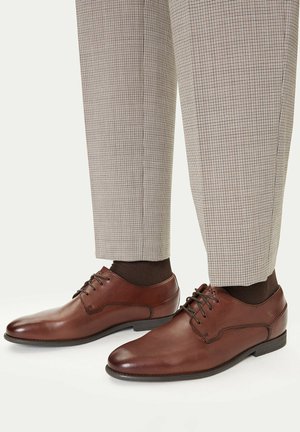 Zapatos de vestir de cuero marrón con cordones, usados con calcetines oscuros y pantalones a medida beige con cuadros.