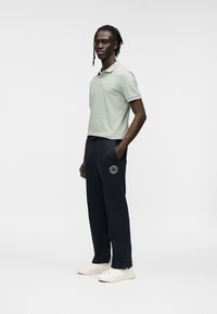 Ανοιχτό πράσινο κοντομάνικο πόλο πουκάμισο σε συνδυασμό με ναυτικό μπλε παντελόνι φόρμας που έχει κυκλικό λογότυπο στον μηρό και λευκά αθλητικά παπούτσια.