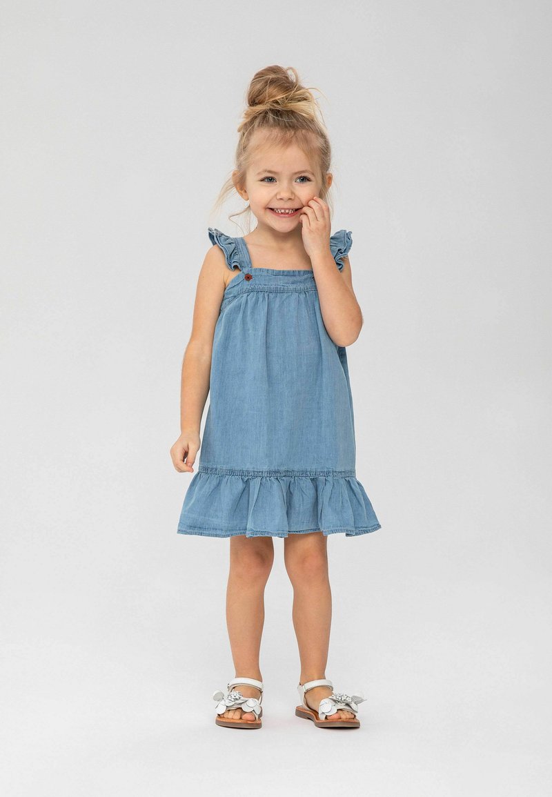 Robe en denim avec des bretelles à volants, une jupe évasée avec un ourlet à volants et un petit bouton rouge en accent. Sandales avec des embellissements floraux.