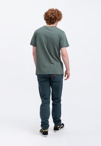 Groene ronde hals t-shirt met korte mouwen, gecombineerd met turquoise broek. De broek heeft een rode accent op de achterzak. Zwarte schoenen met witte zolen.