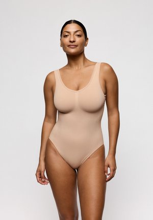 Vrouw met een middelhuidkleur en donker haar in een staart, gekleed in een naadloos nude mouwloos bodysuit, staande tegen een effen lichte achtergrond.