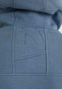 Modrý svetrový materiál s ribovanou texturou. Obsahuje vyšívané logo a výrazné švy na panelech. S kapucí a velkou kapsou.