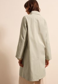 Lichtgroene, middellange jas van glad textiel. Heeft een klassieke kraag, lange mouwen en zij-splitten voor meer bewegingsvrijheid.