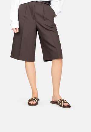 Persona que lleva pantalones cortos a medida de color marrón oscuro hasta la rodilla, camiseta blanca de manga larga y sandalias marrones abiertas con tiras trenzadas.