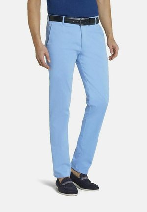 Mann trägt hellblaue, gerade geschnittene Hosen, schwarzen Gürtel, marineblaue Loafers und ein in die Hose gestecktes blaues Hemd, steht vor einem weißen Hintergrund.