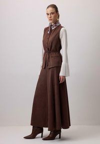 Obleka iz rjavega semiša s prilegajočim se zgornjim delom, polno krilo in gumbi. Bela majica z dolgimi rokavi z naborki. Vključuje vzorčast šal in škornje z visokimi petami.