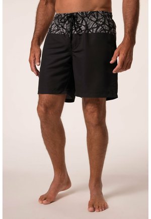 Hombre con bañador negro con una cinturilla estampada, de pie descalzo sobre un fondo neutro.