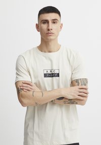 Kremfarget t-skjorte med korte ermer og et svart rektangulært grafisk motiv som leser "A.R.C." sammen med japanske tegn. Laget av bomull, slim fit, med rund hals.