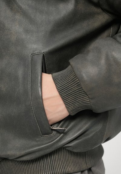 Pilka odinė bombonešio striukė turi elastines rankogalius, kišenę su užtrauktuku ir tekstūruotą paviršių, akcentuojant jos struktūruotą dizainą ir medžiagą.