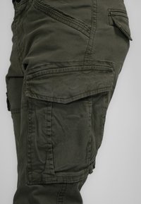 Cargo pants in dark olive green fabric, featuring multiple pockets, visible stitching, and a slightly distressed texture.