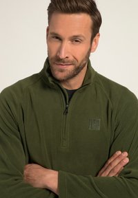 Pullover in pile verde oliva con zip quart, caratterizzato da una discreta patch con logo e cuciture piatte. Tessuto morbido e maniche lunghe.