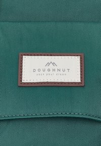 Tissu vert avec une pièce rectangulaire blanche cousue bordée de marron, affichant le logo de la marque Doughnut et le slogan « pack your dream ».