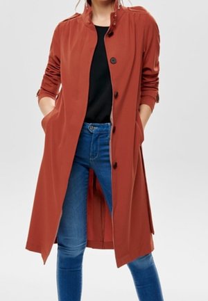 Vrouw draagt een lange, roestkleurige jas met knopen over een zwart shirt en blauwe spijkerbroek, met handen in de zakken van de jas.