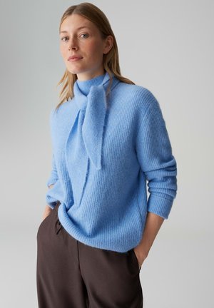 Lichtblauwe gebreide trui met een hoge kraag en een strikdetail aan de voorkant. Heeft lange mouwen en een gestructureerde afwerking. Gecombineerd met donkere broek.