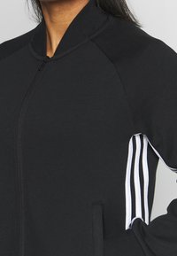 Musta vetoketjullinen urheiluquicki, jossa on pystykaulus, raglan-hihat ja valkoiset kolmiraitasiritykset sivuilla. Pehmeä kangasmateriaali.