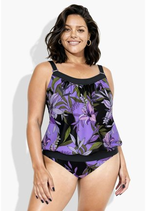 Donna che indossa un tankini floreale viola e nero con orecchini a cerchio dorati, sorridente, su sfondo bianco semplice.