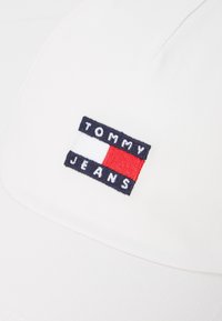 Biely klobúk s látkovou nášivkou loga s nápisom "TOMMY JEANS" v námorníckej farbe a červeno-bielym obdĺžnikovým dizajnom. Mäkká textúra, jednoduchý dizajn.