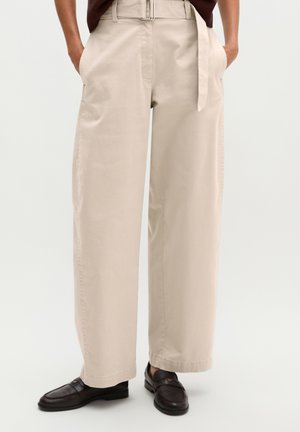 Pantalon beige à jambes larges avec passants de ceinture et ceinture en tissu, porté avec des mocassins marron foncé et les mains dans les poches.