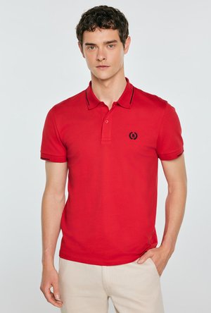 Młody mężczyzna w czerwonej koszulce polo z krótkim rękawem z czarnym haftowanym logo i beżowych spodniach, stojący na jasnym, jednolitym tle.