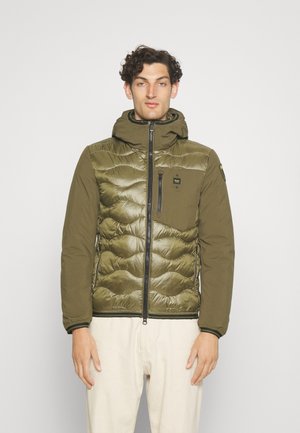 Giacca imbottita verde oliva con cappuccio, caratterizzata da una chiusura frontale con zip, design in tessuto trapuntato e zip laterali. Accentuata da dettagli in metallo nero.
