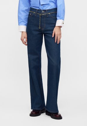 Persona che indossa jeans blu scuro a gamba dritta con una cintura decorativa a catena dorata, abbinata a una camicia blu e mocassini scuri.