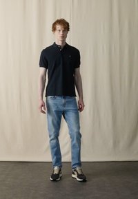 Granatowa koszulka polo, jasnoniebieskie dżinsy, czarne i szare trampki. Model stoi na beżowym tle. Koszulka polo ma kołnierzyk i logo.