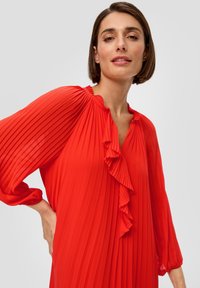 Vrouw met kort bruin haar, gekleed in een helderrode geplooide blouse met ruches, poserend tegen een effen lichte achtergrond.