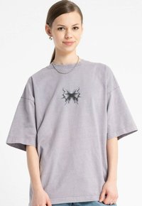 Młoda kobieta z włosami związanymi w kok nosi oversize'owy, wyblakły fioletowy T-shirt z czarnym abstrakcyjnym nadrukiem i srebrny naszyjnik, delikatnie się uśmiechając.