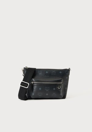 Sac bandoulière noir avec sangle réglable, poche frontale zippée argentée et motif discret du logo MCM sur cuir texturé, sur fond blanc.