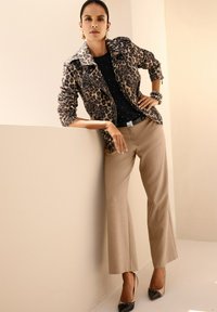 Vrouw leunend tegen een beige muur, gekleed in een jas met luipaardprint, een zwarte top, beige broek, zwarte puntige hakken en gouden creolen.