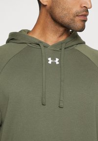 Olivgrüner Kapuzenpullover mit einer vorderen Kängurutasche, Zugkordelverschluss und weißem Under Armour-Logo auf der Brust.