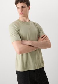 Lysegrøn kortærmet t-shirt lavet af blød bomuld, med rund hals og et lille hvidt logo på brystet, sammen med mørke jeans.