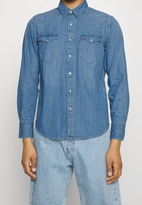 Blå denimskjorta med lång ärm, spetsig krage, tryckknappar, två bröstfickor och en lätt rundad nederkant. Bärs med ljusa denimbyxor.