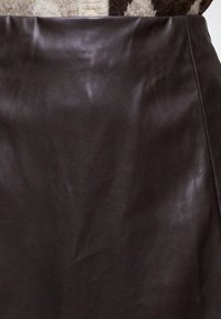 Jupe en cuir marron foncé à texture lisse, suspendue devant un fond avec un tissu en laine à motifs et un bouton blanc.
