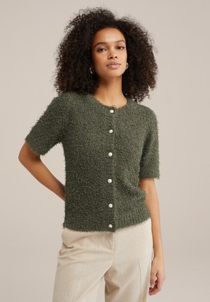 Green textured short-sleeve cardigan with rounded neckline and white button closures, paired with light-colored tailored pants.