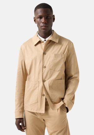 Mand iført beige knapjakke med tre forlommer og matchende bukser, stående med den ene hånd i lommen.