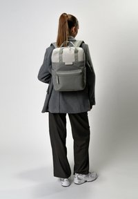 Zaino grigio e crema con più tasche, materiale in tessuto, manico resistente e spallacci regolabili, indossato sulla schiena.