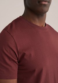 Bordeauxrode katoenen T-shirt met korte mouwen, ronde hals, gladde textuur en subtiele stikseldetails aan de zoom en mouwen.