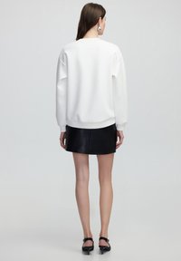 Sudadera blanca con hombros caídos, puños acanalados y cuello redondo, combinada con una falda corta de cuero negro y tacones negros con tiras.