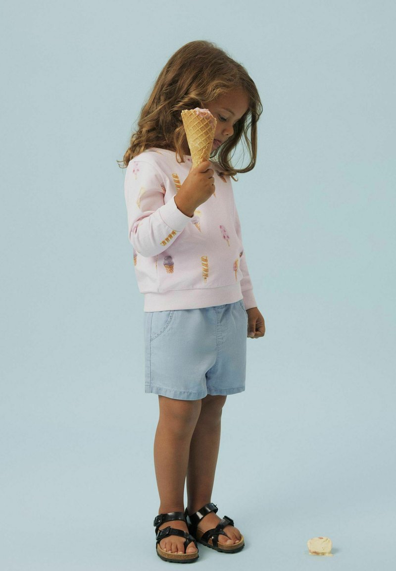 Niño con sudadera de un rosa claro con patrones de conos de helado, combinado con pantalones cortos azules claros y sandalias negras, sosteniendo un cono de helado.