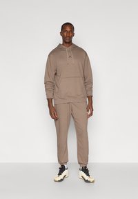 Hnedá mikina s predným vreckom a kapucňou na šnúrku, spojená s zodpovedajúcimi zúženými teplákmi a bielymi teniskami s čiernymi akcentmi.