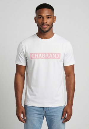 Hombre con camiseta blanca con un logo rectangular rosa que dice "CHABRAND" y jeans azul claro, de pie contra un fondo gris.