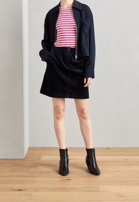 Chaqueta recortada azul marino sobre una camiseta de rayas rosa y blanca, combinada con una falda de pana negra y botas negras hasta el tobillo. Fondo de suelo de madera.