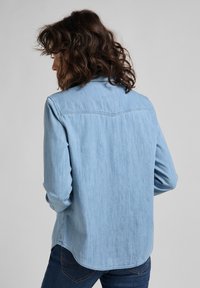 Donna con capelli ricci e scuri, che indossa una camicia di jeans blu chiaro e jeans scuri, girata di spalle con le mani nelle tasche su uno sfondo semplice.