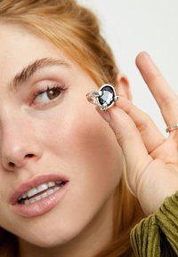 Bague en argent ornée d'une grande pierre précieuse noire facettée, tenue par une main sur un fond neutre, avec des détails complexes sur le bracelet.