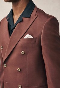 Blazer burdeos de doble botonadura hecho de tejido texturizado, con grandes botones crema y un cuello de camisa negra debajo. Detalle de pañuelo blanco en el bolsillo.