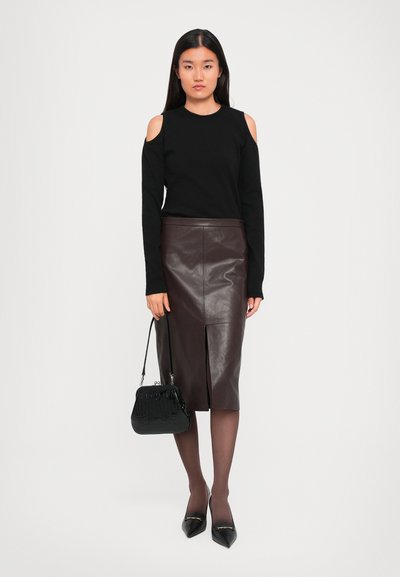 Woman wearing a black cold-shoulder sweater, brown leather knee-length skirt, sheer tights, black pointed heels, holding a black handbag.