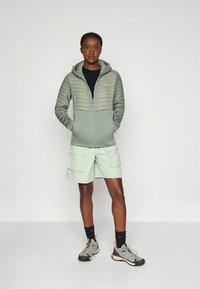 Svetlo zelena podložena jakna z zadrgo, kombinirana s svetlimi kratkimi hlačami. Zgornji del ima teksturo, spodnji del je enobarven, kapuca je oprijeta.